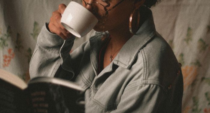 brown skinned woman with curly hair drinkig out of a white cup while reading a book shes wearing glasses and a denim jacket.jpg.optimal