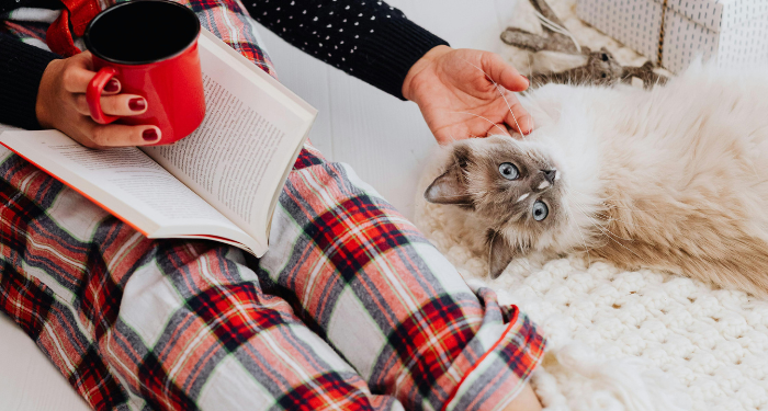 person in plaid pjs petting cat while holding a mug and a book in their lap